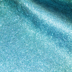 May include: Close-up of a textured, shimmering teal surface. The material has a subtle, granular pattern with highlights that catch the light, creating a sparkling effect. The overall appearance is smooth and luxurious.