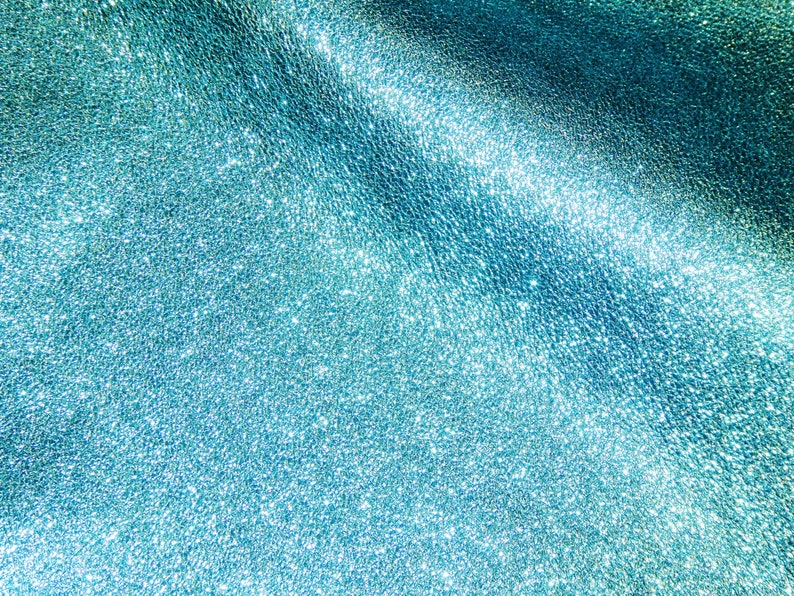 May include: Close-up of a textured, shimmering teal surface. The material has a subtle, granular pattern with highlights that catch the light, creating a sparkling effect. The overall appearance is smooth and luxurious.