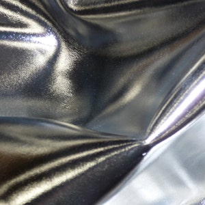 May include: Close-up of a silver metallic fabric with a smooth, shiny surface. The fabric is folded and draped, creating soft, flowing lines and folds.