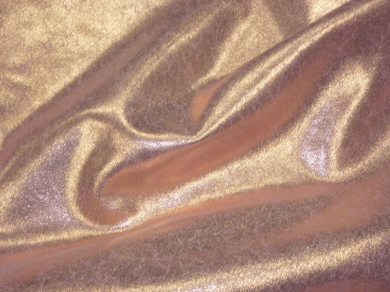 K&ouml;nnte beinhalten: Nahaufnahme eines glatten, metallischen Stoffes mit einem subtilen Schimmer. Der Stoff ist hellgoldfarben und hat eine weiche, flie&szlig;ende Textur.