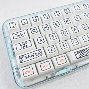 Könnte beinhalten: Nahaufnahme einer weißen Tastatur mit schwarzer und orangefarbener Beschriftung. Die Tasten sind in einem QWERTZ-Layout angeordnet, mit Tasten mit der Aufschrift "Tab", "Shift", "Ctrl", "Win" und "Alt". Die Tastatur ist in einem transparenten, hellblauen Rahmen eingefasst.