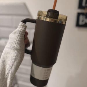 Puede incluir: Un vaso t&eacute;rmico de color marr&oacute;n oscuro con asa a juego y tapa transl&uacute;cida de color &aacute;mbar. El vaso presenta una base delgada y c&oacute;nica e incluye una pajita naranja recta insertada a trav&eacute;s del centro de la tapa para poder beber.