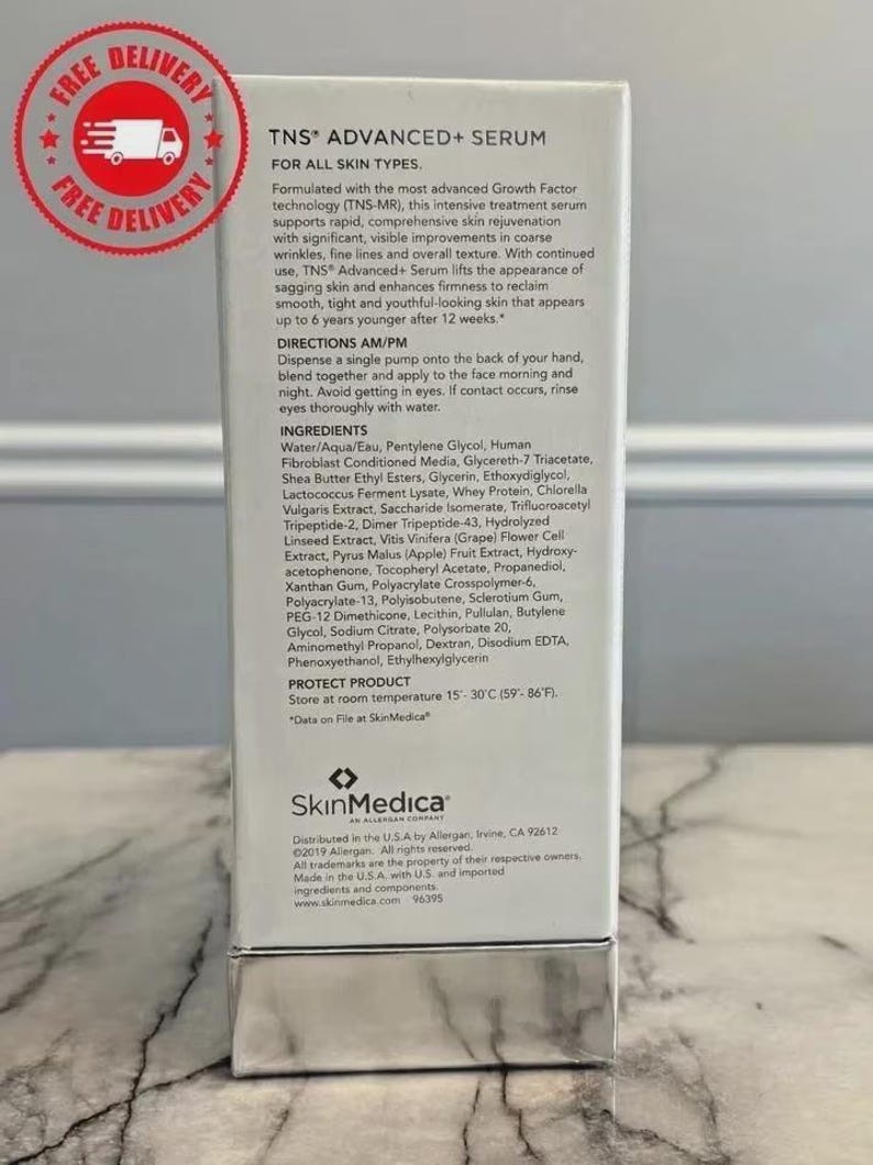 May include: A white rectangular box of SkinMedica TNS Advanced+ Serum. The front features black text detailing product benefits, directions, and ingredients. A silver metallic base is at the bottom. A red circular free delivery stamp is in the corner.