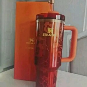May include: A red translucent Stanley tumbler with an orange handle and a matching orange lid. The cup features a floral pattern and the Stanley logo in gold. A gold-colored straw topper shaped like a pineapple is attached to the straw.