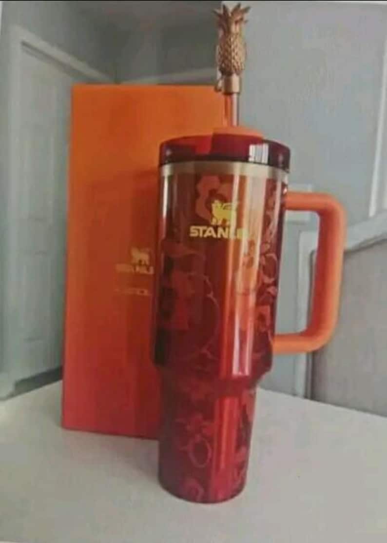 May include: A red translucent Stanley tumbler with an orange handle and a matching orange lid. The cup features a floral pattern and the Stanley logo in gold. A gold-colored straw topper shaped like a pineapple is attached to the straw.