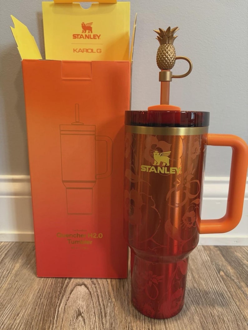May include: A metallic orange Stanley Quencher H2.0 tumbler featuring a floral pattern, a gold-toned pineapple straw topper, and an orange handle. The tumbler is displayed next to its matching orange and yellow packaging box labeled Stanley and Karol G.