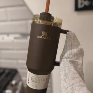 Puede incluir: Un vaso Stanley Quencher H2.0 FlowState de color marr&oacute;n oscuro con asa a juego, tapa transparente y pajita marr&oacute;n. El vaso presenta el logotipo dorado de Stanley y una etiqueta blanca cerca de la base que dice Quencher H2.0 Tumbler.