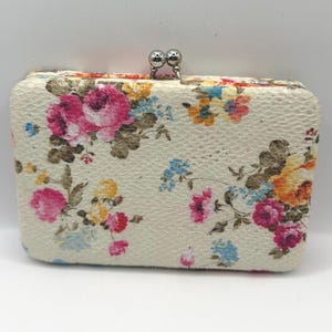 May include: A rectangular clutch with a floral pattern featuring pink, yellow, and blue flowers on a cream background. The clutch has a silver clasp closure.
