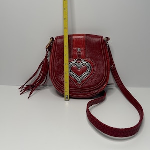 Op de afbeelding: Een rode leren crossbodytas met een zilveren hartvormige versiering. De tas heeft een gevlochten riem en een kwast. De tas is ongeveer 25 cm hoog.
