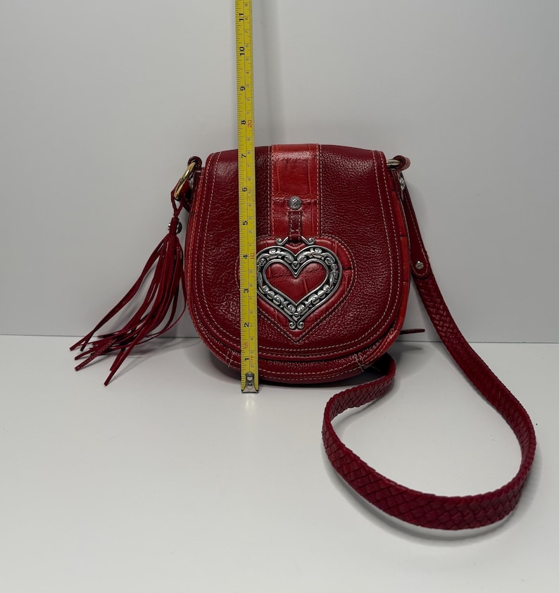 Op de afbeelding: Een rode leren crossbodytas met een zilveren hartvormige versiering. De tas heeft een gevlochten riem en een kwast. De tas is ongeveer 25 cm hoog.