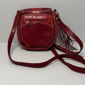 Op de afbeelding: Rode leren crossbodytas met een gevlochten riem en een klepsluiting. De tas heeft een getextureerd voorpaneel, een vak met ritssluiting en decoratieve franjes. De stiksels zijn wit en de hardware is goudkleurig.