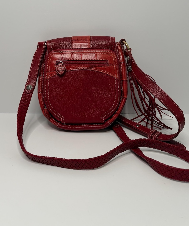 Op de afbeelding: Rode leren crossbodytas met een gevlochten riem en een klepsluiting. De tas heeft een getextureerd voorpaneel, een vak met ritssluiting en decoratieve franjes. De stiksels zijn wit en de hardware is goudkleurig.