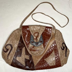 May include: A vintage shoulder bag with a patchwork design in shades of brown and tan. The bag features a textured, reptile-like pattern and a decorative silver-tone clasp. The long strap is made of matching material.