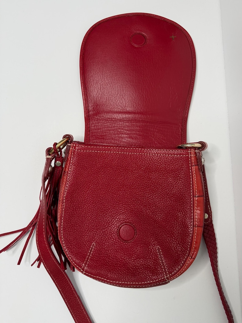 Op de afbeelding: Rode leren zadeltas met klepsluiting, witte stiksels en een lange riem. De tas heeft een textuuroppervlak, een ronde sluiting en decoratieve franjes aan de zijkant. De binnenkant is ook rood.