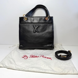 May include: A black leather handbag featuring a centered embossed X logo, gold-tone hardware rings, and dual top handles. The bag is accompanied by a detachable black leather shoulder strap and a white fabric dust bag with the text By Paloma Picasso.