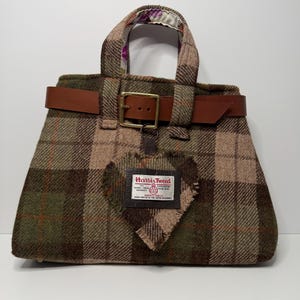 May include: A Harris Tweed handbag with a brown leather belt and buckle closure. The bag features a heart-shaped patch and a label with text. The bag has a plaid pattern in shades of green, brown, and tan.