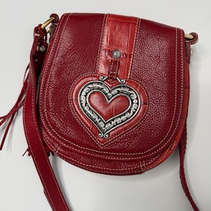 Op de afbeelding: Rode leren crossbodytas met een zilverkleurige hartvormige versiering. De tas heeft een getextureerde leren body, een klepsluiting en een lange verstelbare riem. Het hart is gedetailleerd met een sierlijk ontwerp.