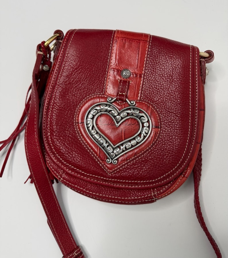 Op de afbeelding: Rode leren crossbodytas met een zilverkleurige hartvormige versiering. De tas heeft een getextureerde leren body, een klepsluiting en een lange verstelbare riem. Het hart is gedetailleerd met een sierlijk ontwerp.