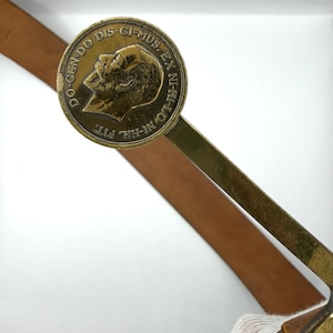 Puede incluir: Hebilla de cinturón de latón vintage con un diseño circular que presenta un retrato de perfil y texto en latín. La hebilla está sujeta a un cinturón de cuero marrón, mostrando una estética clásica e histórica.