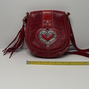 Op de afbeelding: Rode leren crossbodytas met een zilverkleurige hartvormige versiering. De tas heeft een gevlochten riem, een klepsluiting en een kwastdetail. De tas is ongeveer 20 cm breed.