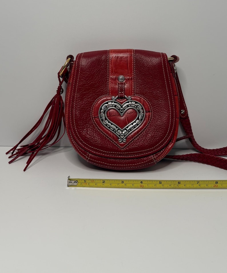 Op de afbeelding: Rode leren crossbodytas met een zilverkleurige hartvormige versiering. De tas heeft een gevlochten riem, een klepsluiting en een kwastdetail. De tas is ongeveer 20 cm breed.