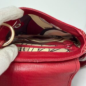 Op de afbeelding: Een close-up van een rode leren handtas met een goudkleurige metalen ring. De binnenkant heeft een voering van stof met een patroon en een zak met ritssluiting. De tas is open en toont de binnenkant.