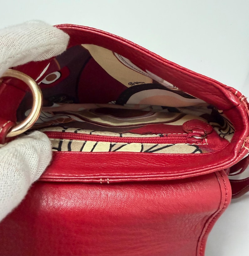 Op de afbeelding: Een close-up van een rode leren handtas met een goudkleurige metalen ring. De binnenkant heeft een voering van stof met een patroon en een zak met ritssluiting. De tas is open en toont de binnenkant.