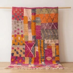 May include: A vibrant, rectangular rug with a patchwork design. The rug displays a mix of pink, orange, yellow, grey, and green geometric patterns. It is hanging on a wooden rod, with a wooden ladder in the background.