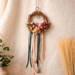 May include: A handcrafted floral wreath with a woven twig base, adorned with dried flowers in pink, yellow, and burgundy hues. Green, pink, and cream ribbons hang below, with small gold bells. A decorative piece for home decor.