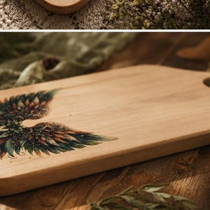 May include: A light-colored wooden cutting board with a decorative wing design. The wings are detailed with shades of green, brown, and orange. The board has a rounded shape with a hole for hanging.