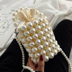 Op de afbeelding: Een crèmekleurige, met parels bezette handtas met een parelachtige buitenkant. De tas heeft een trekkoordsluiting en een met parels bezette handgreep. Er is ook een met parels bezette riem bevestigd. De tas wordt vastgehouden door een persoon met een rode manicure.