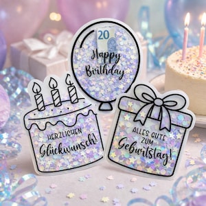 Peut inclure: Trois décorations en acrylique transparent remplies de confettis irisés en forme d'étoiles et de cercles. Un ballon avec 'Happy Birthday' et un billet de 20 euros, un gâteau avec 'HERZLICHEN Glückwunsch!' et un cadeau avec 'ALLES GUTE ZUM Geburtstag!'