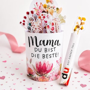 Könnte beinhalten: Ein weißer zylindrischer Behälter mit dem deutschen Text "Mama, DU BIST DIE BESTE!" und floralen Illustrationen. Eine dekorative Blumenanordnung erhebt sich aus dem Behälter. Eine weiße Röhre mit dem Text "BIST EINFACH TOLL" und ein Schokoriegel sind ebenfalls vorhanden.