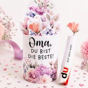 Könnte beinhalten: Eine zylindrische Geschenkbox mit Aquarellblumen und dem deutschen Text "Oma, DU BIST DIE BESTE!" (Oma, du bist die Beste!). Eine Süßwarenriegel mit dem Text "BIST GOLD WERT" (Gold wert) liegt neben der Box. Dekorationen mit Blumen.