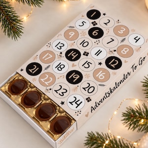 Könnte beinhalten: Ein rechteckiger Adventskalender mit cremefarbener Oberfläche und nummerierten Kreisen von 1 bis 24 in Schwarz, Weiß und Beige. Der Text Adventskalender To Go steht an der Seite. Ein goldenes Fach enthält vier dunkelbraune, ovale Pralinen.