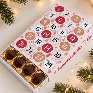 Könnte beinhalten: Ein rechteckiger Adventskalender mit hellrosa Hintergrund und nummerierten Kreisen von 1 bis 24 in Rot, Weiß und Beige. Die Schachtel ist teilweise geöffnet und zeigt eine goldene Kunststoffschale mit mehreren dunkelbraunen Pralinen.
