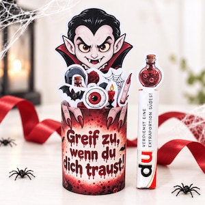 May include: A cylindrical container with a red and black gradient background featuring the text Greif zu, wenn du dich traust. It is decorated with cutouts of a vampire, eyeball, bat, and potion. Beside it stands a white tube labeled du.