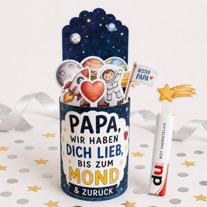 May include: A cylindrical Father's Day card with a space theme. The card features a blue background with planets, stars, and a rocket ship. The text on the card reads "PAPA, WIR HABEN DICH LIEB, BIS ZUM MOND & ZURÜCK". A small flag says "BESTER PAPA". A candy bar says "BIST UNERSETZLICH".