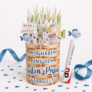 May include: A decorative basket-shaped card with fishing-themed cutouts, including fish, a fishing rod, and a hook. The card has text that reads "WIR HABEN UNS DEN besten Papa GEANGELT!" and "BIST EIN SUPERHELD". Blue ribbon and fish-shaped confetti are scattered around.