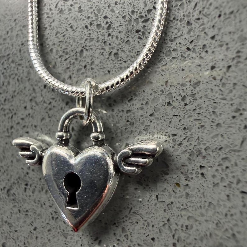 May include: A silver necklace with a heart-shaped padlock pendant, complete with small wings. The pendant, with a keyhole, hangs from a silver chain. The necklace is set against a grey background.