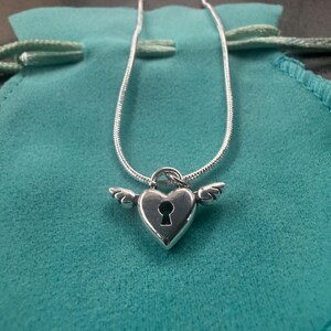 May include: A silver necklace with a heart-shaped pendant, keyhole detail, and wing embellishments. The pendant is suspended from a fine chain. The necklace is displayed on a teal drawstring pouch.