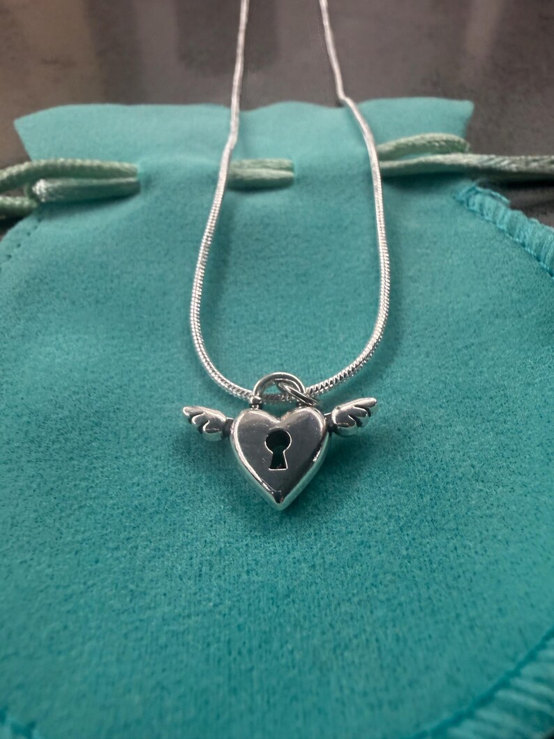 May include: A silver necklace with a heart-shaped pendant, keyhole detail, and wing embellishments. The pendant is suspended from a fine chain. The necklace is displayed on a teal drawstring pouch.