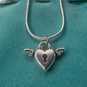 May include: A silver heart-shaped pendant necklace with a keyhole design and small wings on either side. The pendant hangs from a delicate silver chain. The necklace is displayed on a teal-coloured surface.
