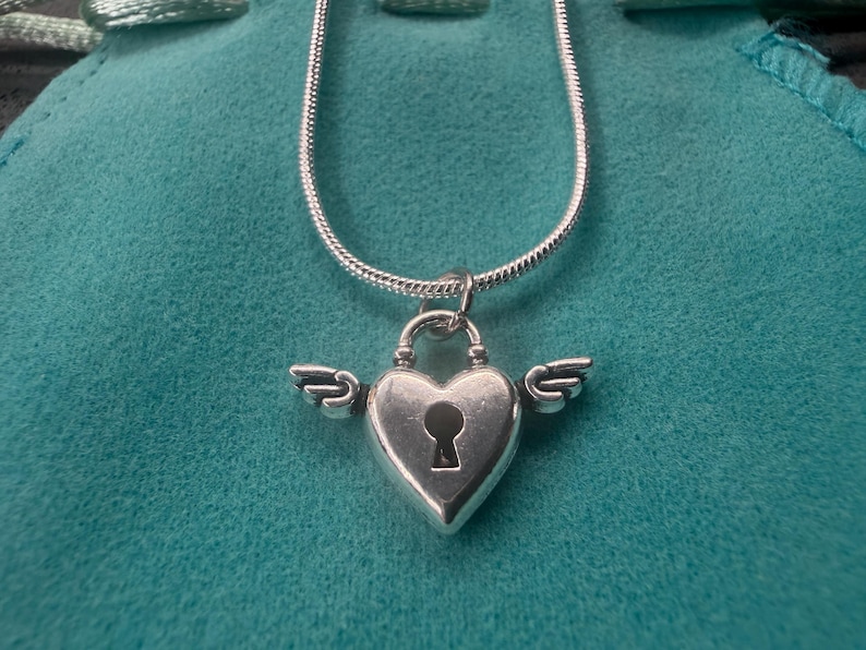 May include: A silver heart-shaped pendant necklace with a keyhole design and small wings on either side. The pendant hangs from a delicate silver chain. The necklace is displayed on a teal-coloured surface.