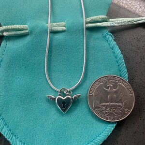 May include: A silver necklace with a heart-shaped pendant, keyhole detail, and wings. The necklace is presented on a teal pouch, with a quarter coin nearby.