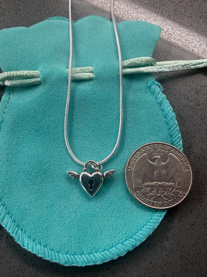 May include: A silver necklace with a heart-shaped pendant, keyhole detail, and wings. The necklace is presented on a teal pouch, with a quarter coin nearby.