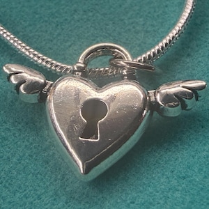 May include: A silver heart-shaped pendant with a keyhole and small wings on a silver chain. The pendant is highly polished and reflects light. The background is a solid teal colour.