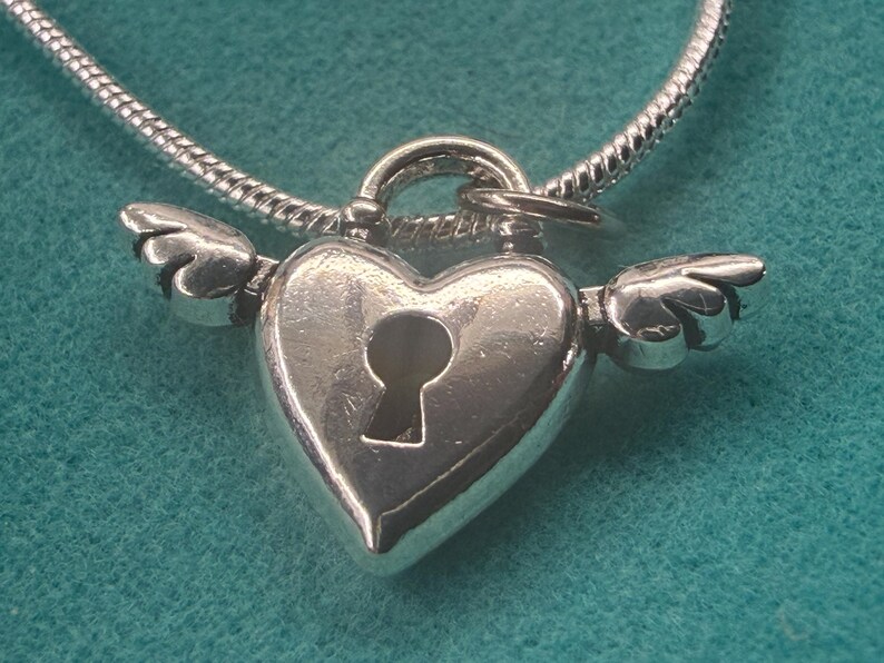 May include: A silver heart-shaped pendant with a keyhole and small wings on a silver chain. The pendant is highly polished and reflects light. The background is a solid teal colour.