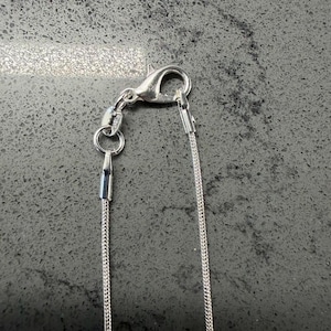 May include: A close-up shot of a silver necklace featuring a lobster clasp and a fine chain. The necklace is set against a dark, textured background. The chain is slender and composed of tiny, linked segments.