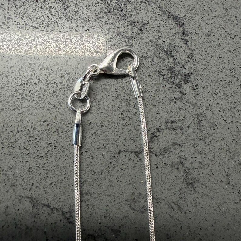 May include: A close-up shot of a silver necklace featuring a lobster clasp and a fine chain. The necklace is set against a dark, textured background. The chain is slender and composed of tiny, linked segments.
